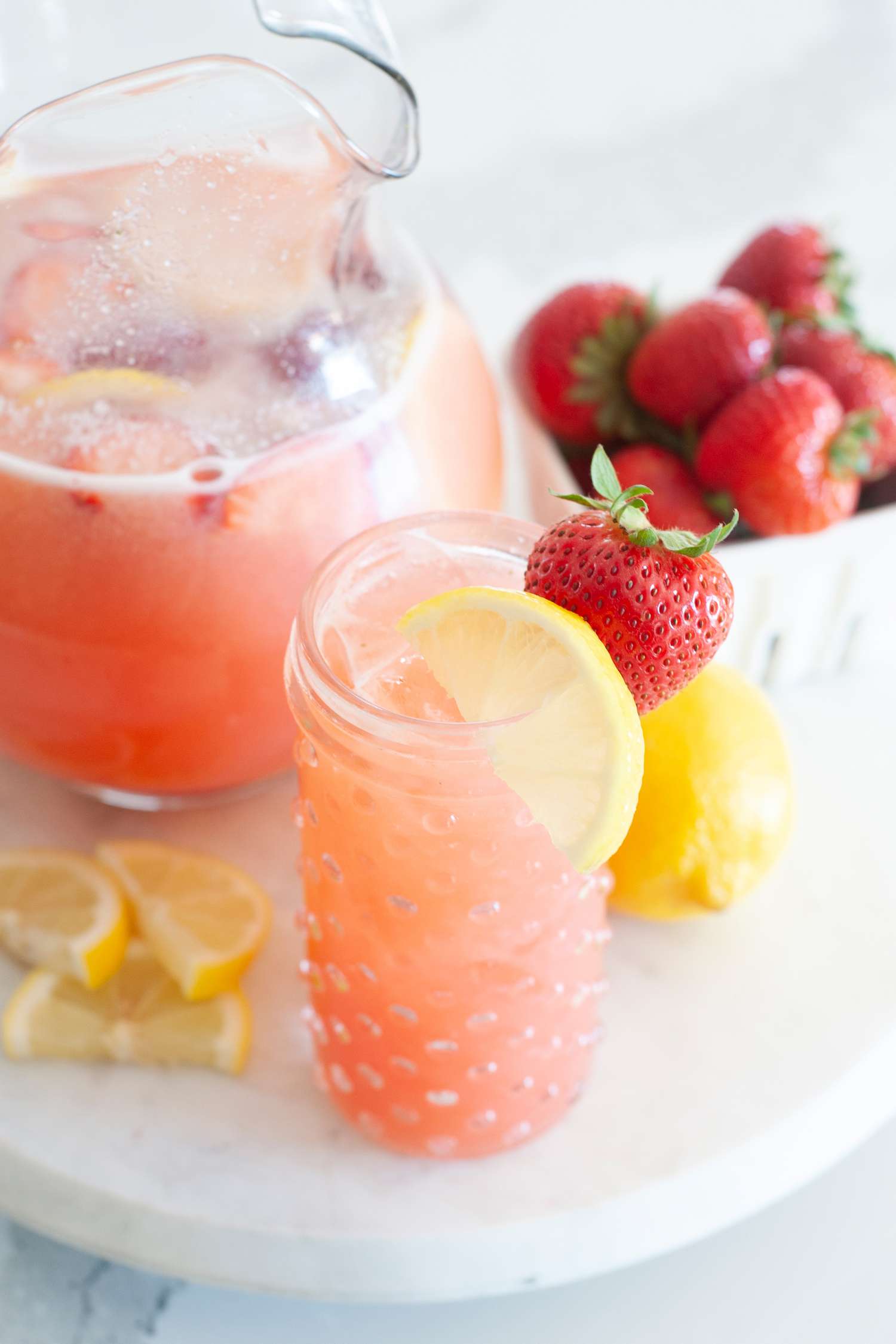 A round platter with a glass and pitcher of blender strawberry lemonade.