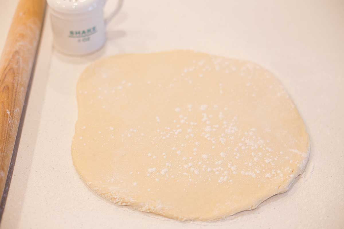 Cafe Du Monde Beignet dough is rolled out and sprinkled with powdered sugar. A rolling pin and sugar shaker are to the left of the dough.