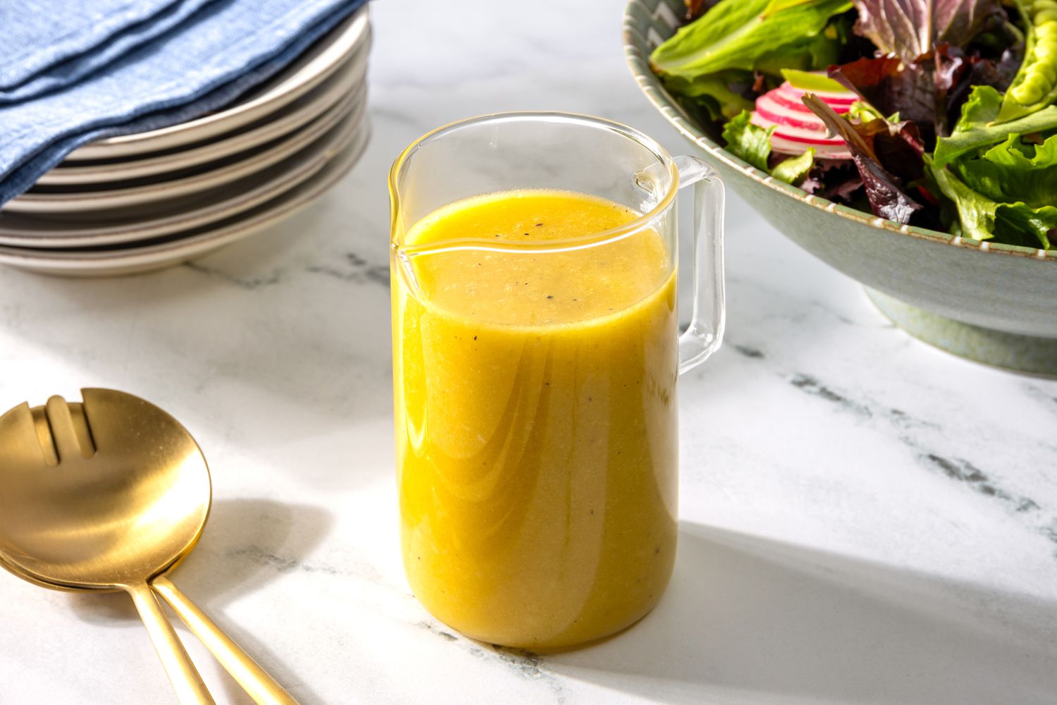 Ginger dressing in a small container at a table setting with a stack of plates, a bowl of salad, and salad utensils