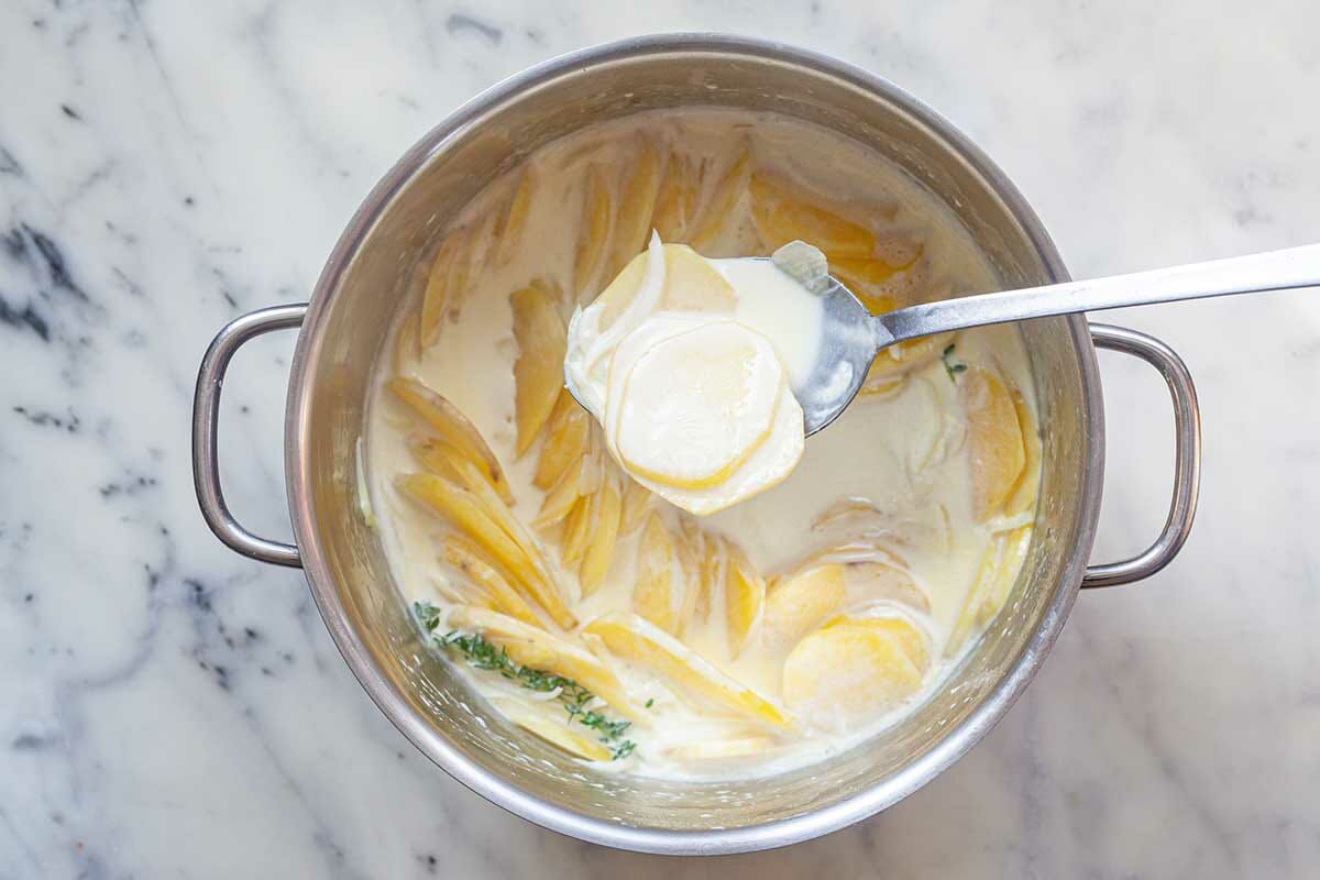 A stock pot has a spoon held above it with a cream sauce covering thinly sliced potatoes to make Creamy Scalloped Potatoes.