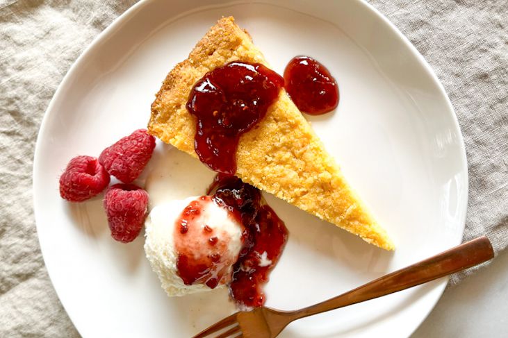 A slice of golden cornmeal cake garnished with raspberry jam, fresh raspberries, and a scoop of ice cream