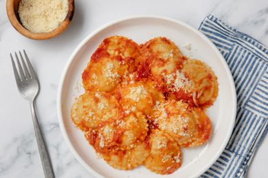 Dish of ravioli with red sauce and grated cheese on a white plate, fork and cloth napkin beside it