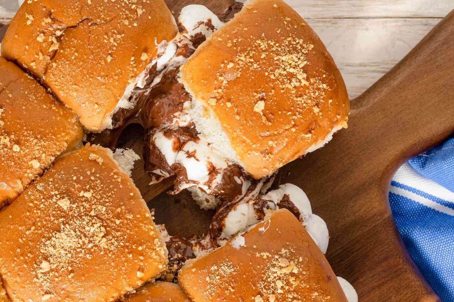 S'mores Sliders Clustered Together on a Wooden Cutting Board With One of the Sliders Ripped Off to Showcase Melted Gooey Chocolate and Marshmallows