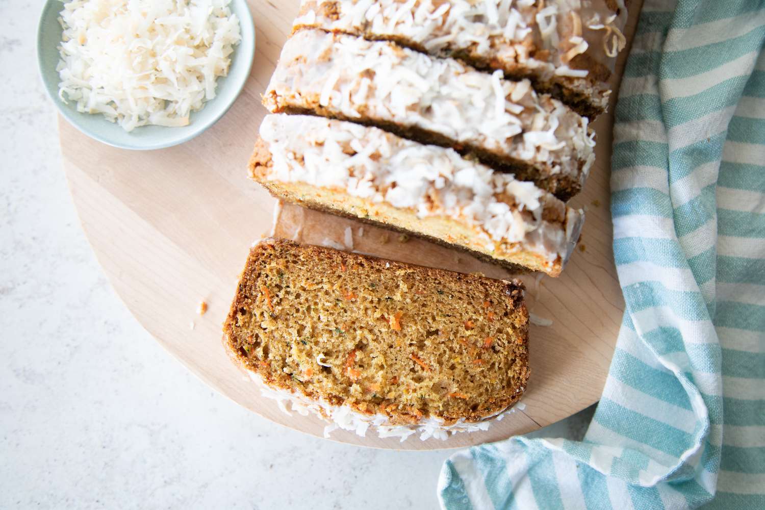 Slices of Zucchini Carrot Bread on Round Wood Board Surrounded by a Kitchen Towel and a Small Bowl of Coconut Flakes