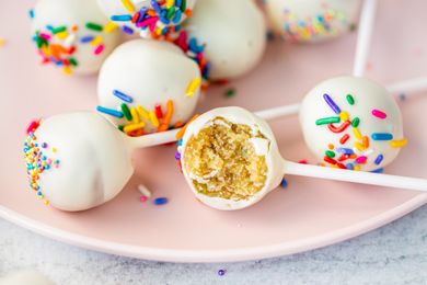 Cake pops coated in a white glaze topped with colorful sprinkles with one revealing its interior on a pale pink plate