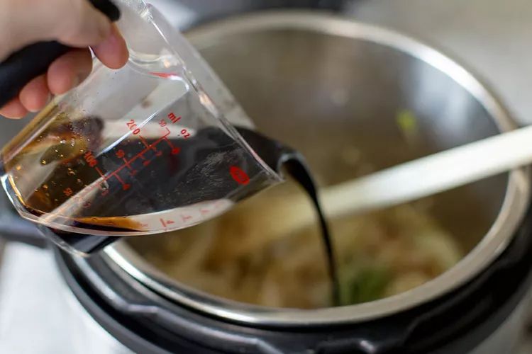 Pouring a dark liquid from a measuring cup into a pot cooking in process