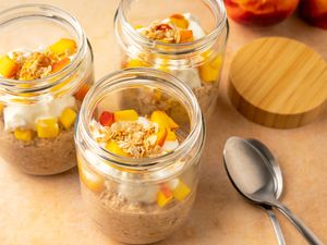 Three jars of peach cobbler overnight oats topped with diced peaches and granola two spoons placed on the table beside them