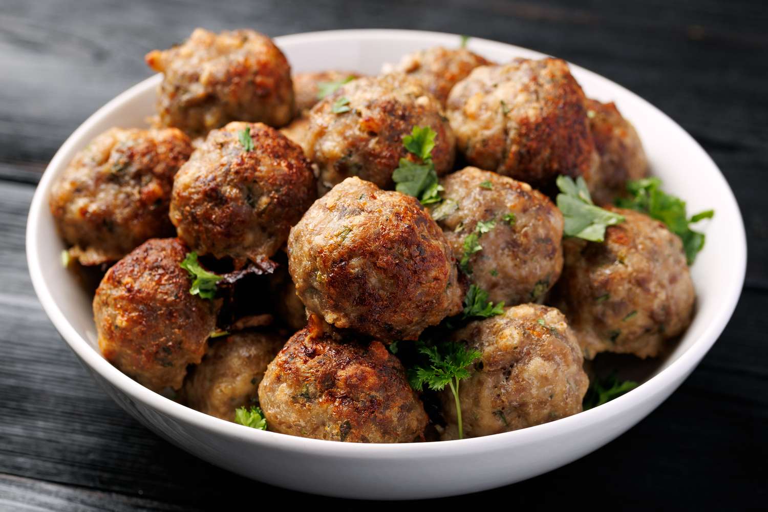 A closeup of a white bowl filled with cooked meatballs garnished with parsley