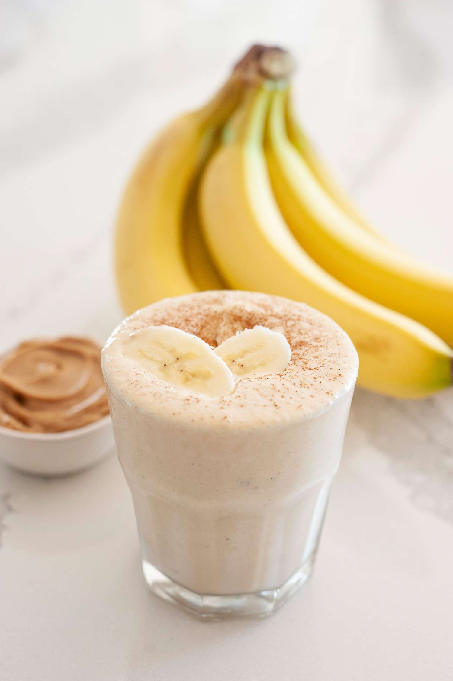 Simple peanut butter banana smoothie topped with sliced bananas and a bunch of bananas and a small container of peanut butter behind it.