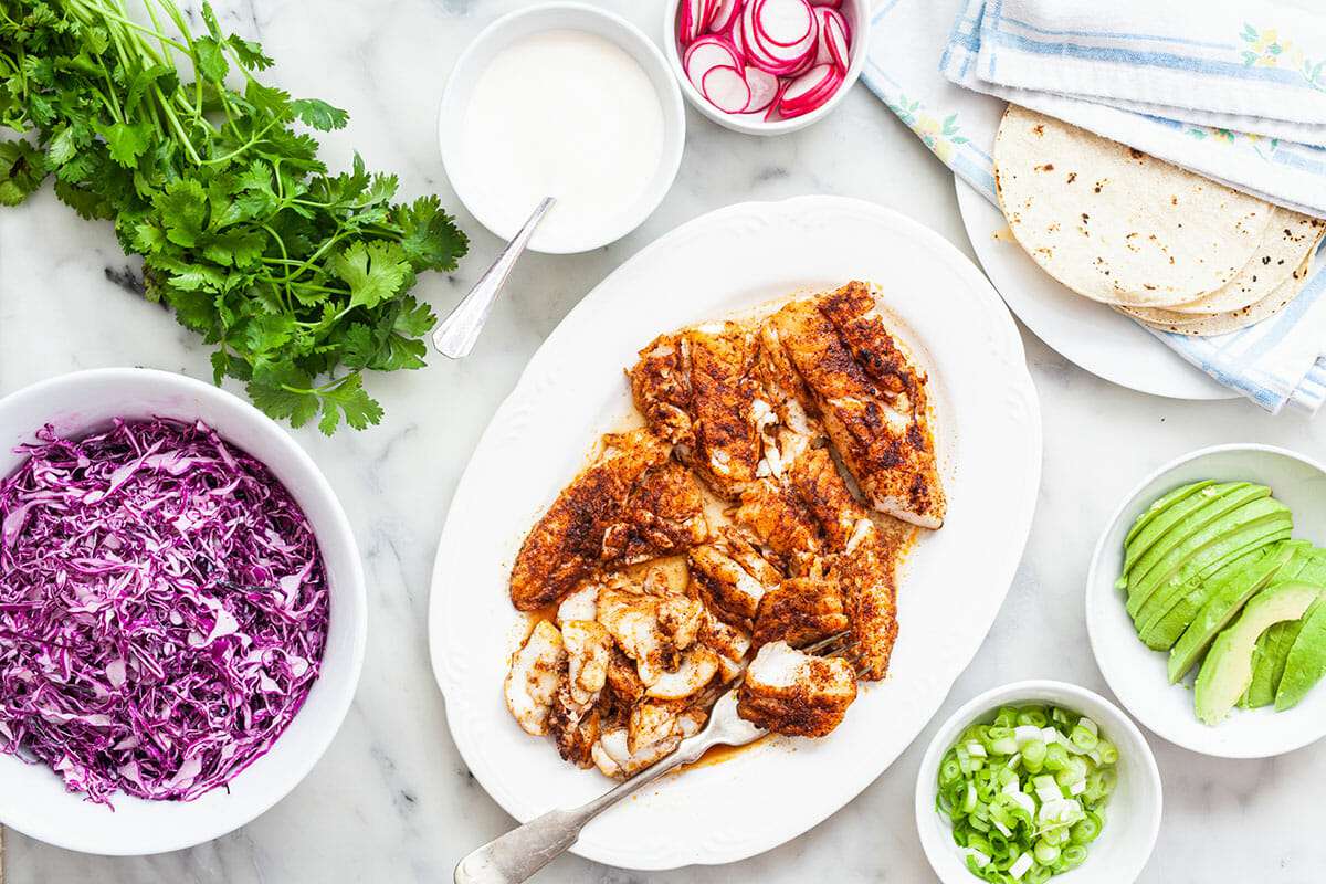 A platter of fish is surrounded by ingredients to top fish tacos to show how to make fish tacos.