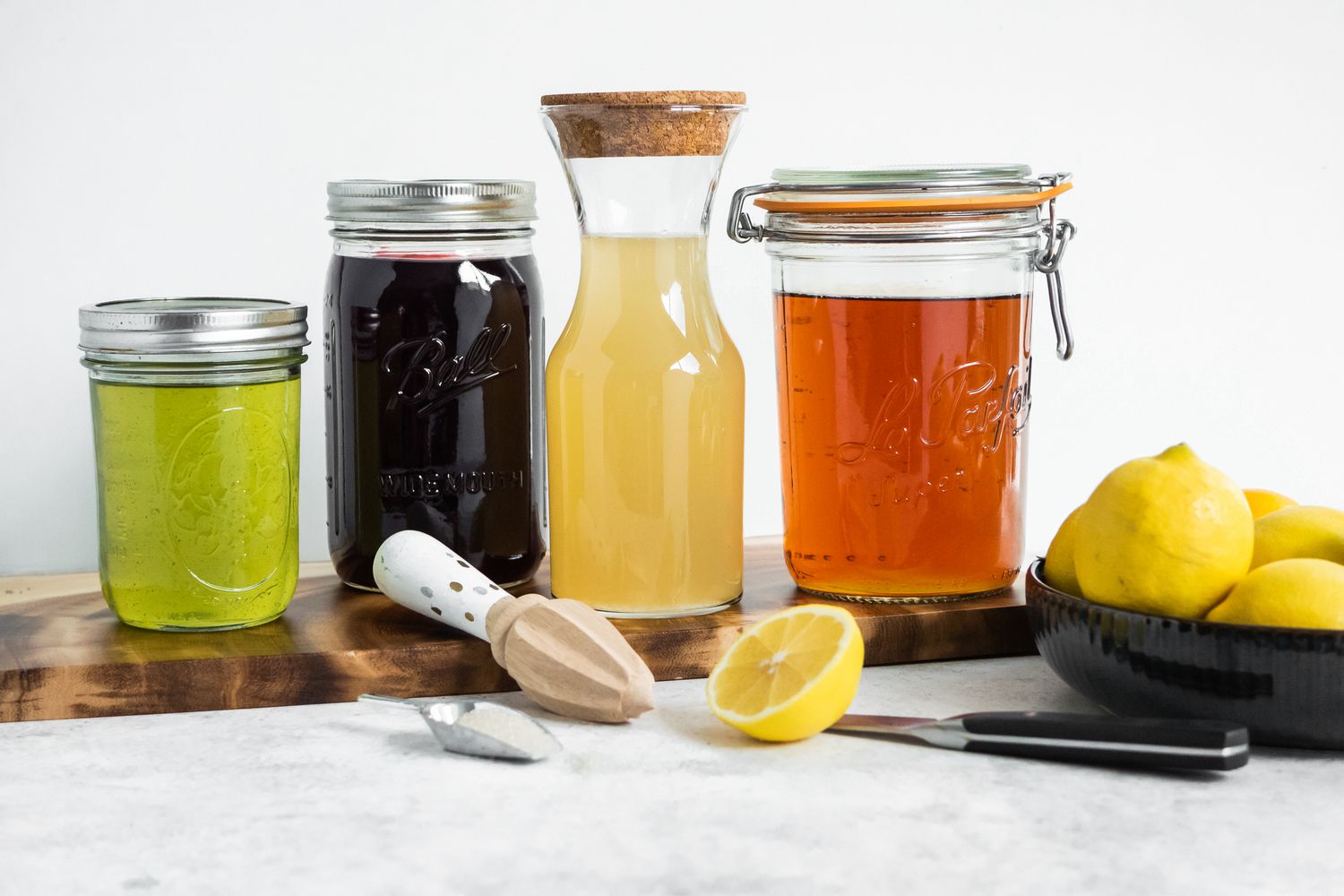 Jars on a Wooden Board (R to L): Jar of Green Tea, Jar of Hibiscus Flower Tea, Jar of Lemon Juice, and a Jar of Black Tea, and in the Surroundings, a Bowl of Lemons, a Knife on the Counter, a Halved Lemon, and a Citrus Reamer