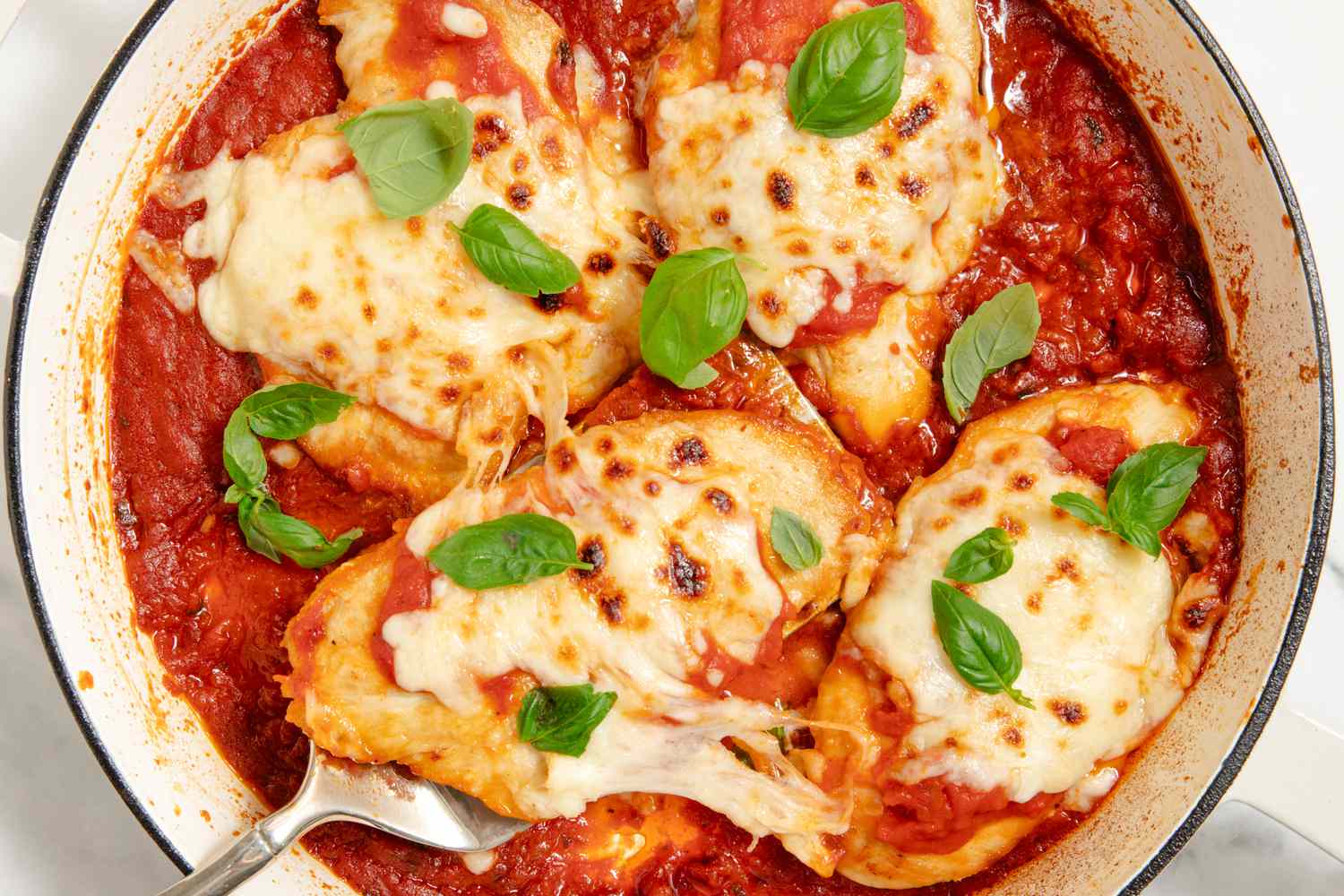 A skillet with four cheese covered chicken breasts in tomato sauce, garnished with basil