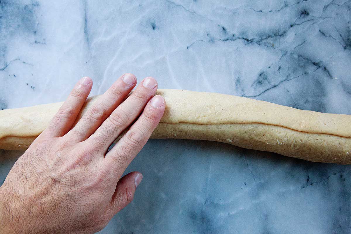 The best King Cake dough is rolled into a tube shape with a hand resting on top. The dough rests on a marble background.