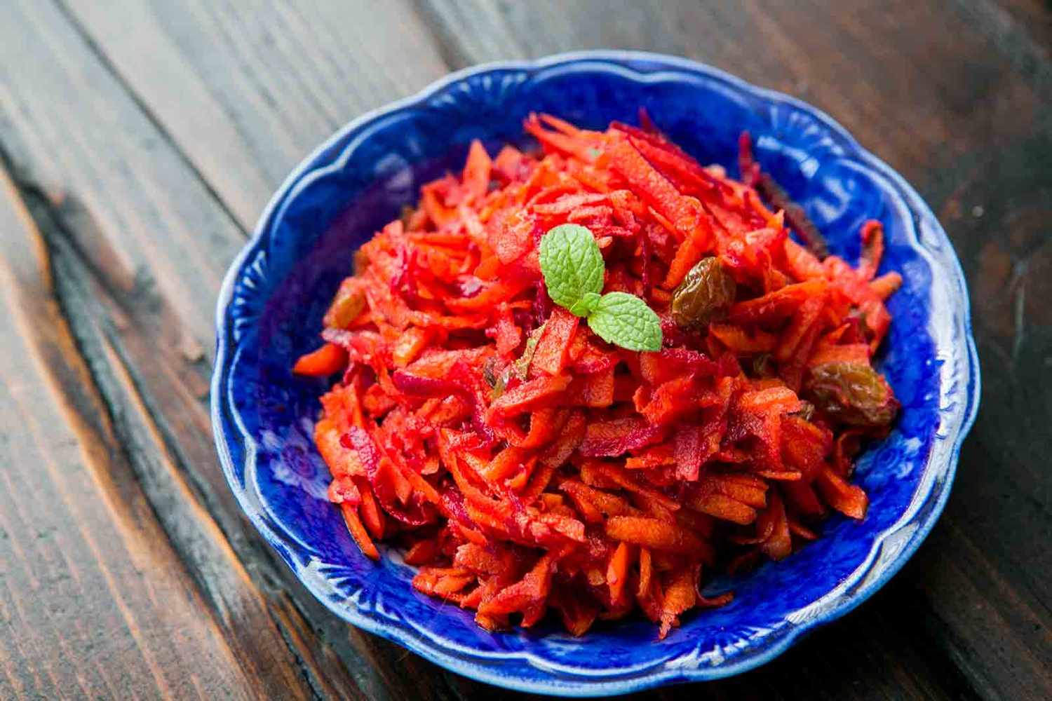A bowl of grated carrot and beet salad garnished with a sprig of mint served in a blue dish on a wooden surface