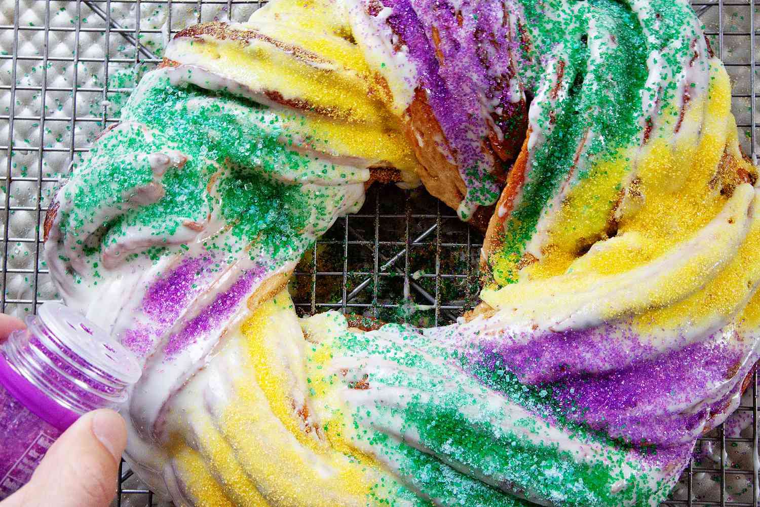 Homemade King Cake on a cooling rack getting topped with purple sprinkles. Yellow, green and purple sprinkles are already on the cake.