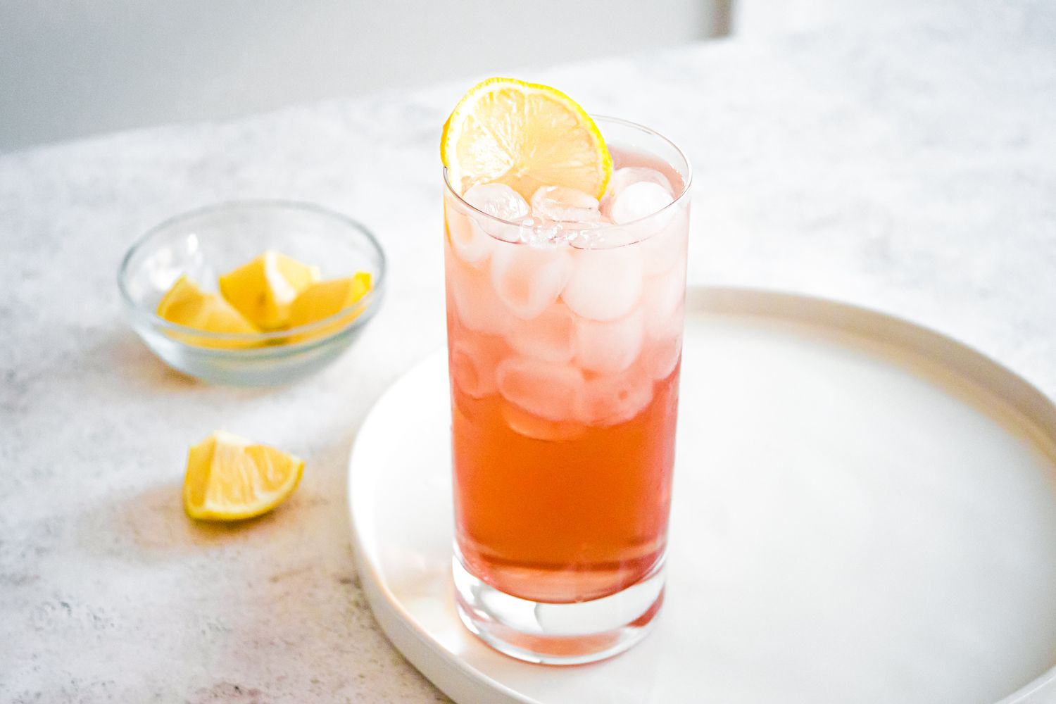 Sloe Gin Fizz in Glass with Ice and Lemon Slice on Tray. Bowl of Lemon Wedges in Background.