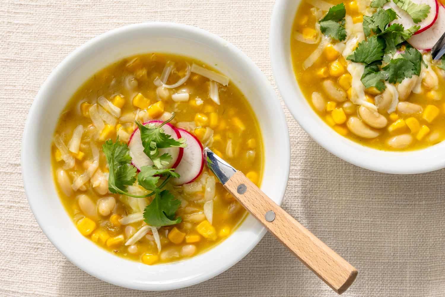 Two bowls of chili verde with corn beans and garnished with radish slices and cilantro a spoon placed on one bowl
