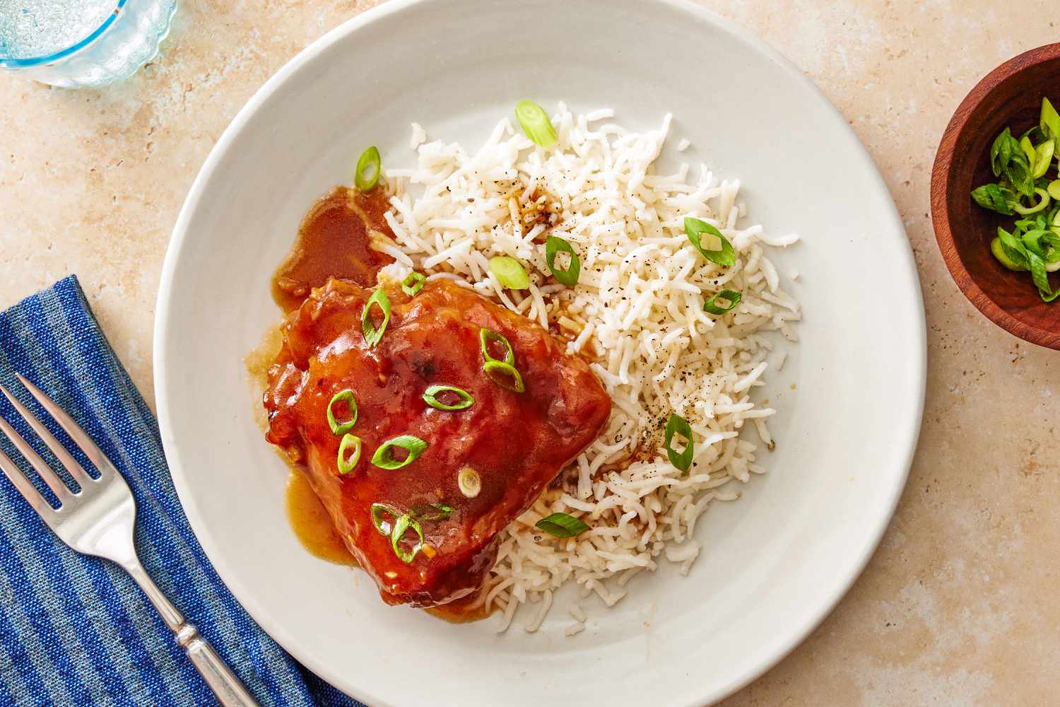 Overhead view of a white plate of soy chicken with rice topped with scallions next to a fork and napkin and a bowl of scallions on a beige background