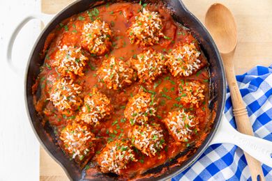 Overhead view of a cast iron skillet of meatballs with expanded rice on the surface and sauce on a wooden cutting board next to a checkered blue and white towel and wooden spoon