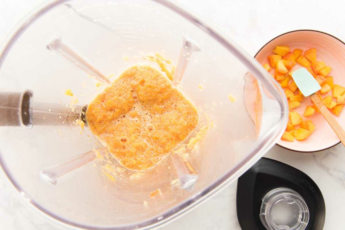 Overhead view of a blender with peach puree inside to make easy peach cobbler ice cream. Chopped peaches are in a bowl to the right.