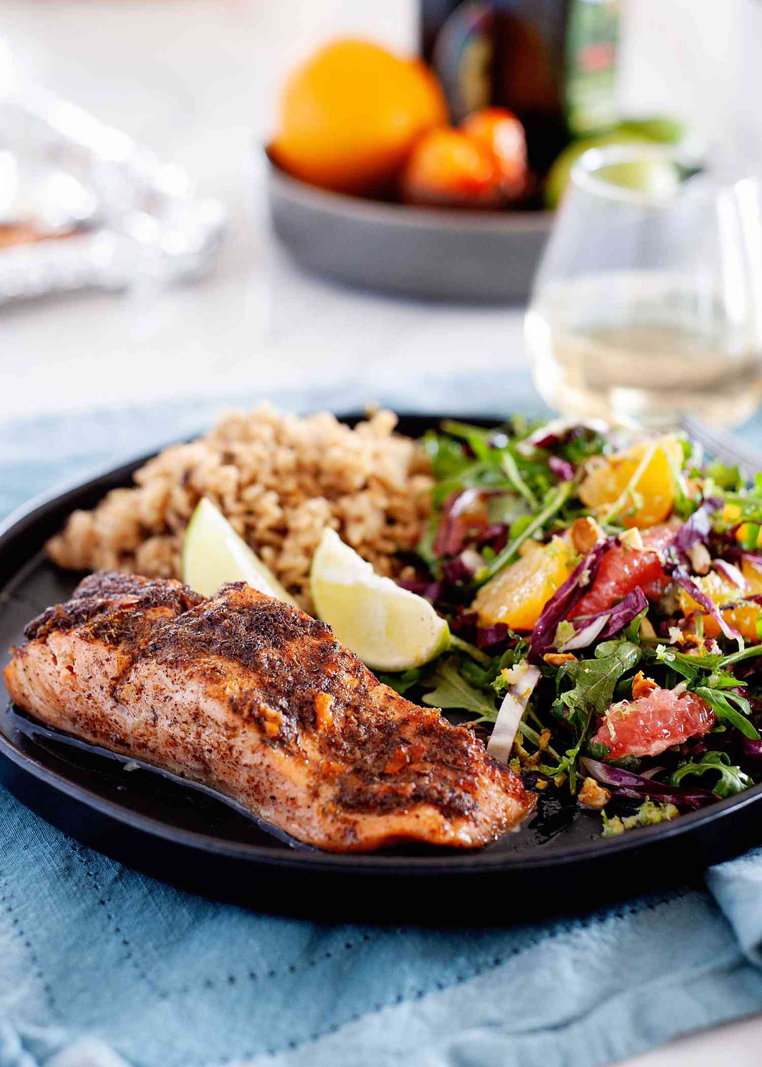 Side view of a fillet of broiled jerk salmon. The salmon has juices pooled at the bottom of it. A mixed green salad with citrus, two lime wedges and rice are visible on the plate behind the salmon. The plate is on a blue linen. Behind the plate is a glass of white wine, cutlery, a bowl of produce and the foil-lined baking sheet.