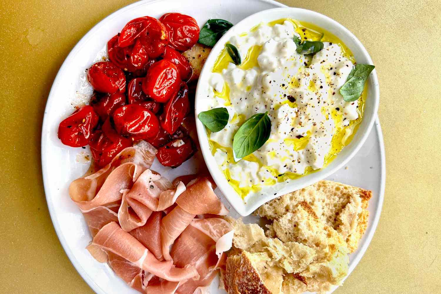 Plate With Cooked Red Cherry Tomatoes, Prosciutto, Bread, and a Bowl of Cottage Cheese Topped With Olive Olive, Basil Leaves, and Cracked Pepper