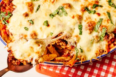A closeup of baked ziti topped with melted cheese and herbs in a dish with a serving spoon lifting a portion from the dish
