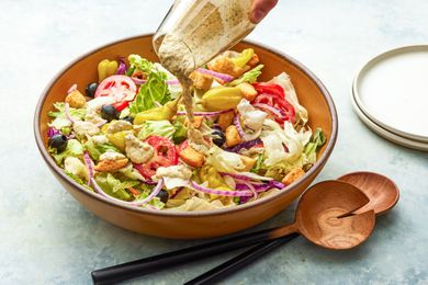 Salad Dressing Poured Over a Bowl of Olive Garden Salad, and on the Counter Next to It, a Stack of Plates and a Set of Salad Serving Utensils