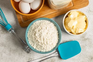 Baking ingredients and tools including flour eggs butter a whisk and a spatula on a countertop