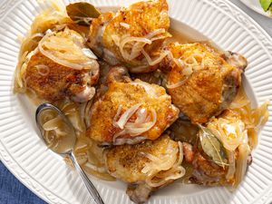 Overhead view of a white platter of six wine roasted chicken thighs topped with onions and bay leaves along with a spoon