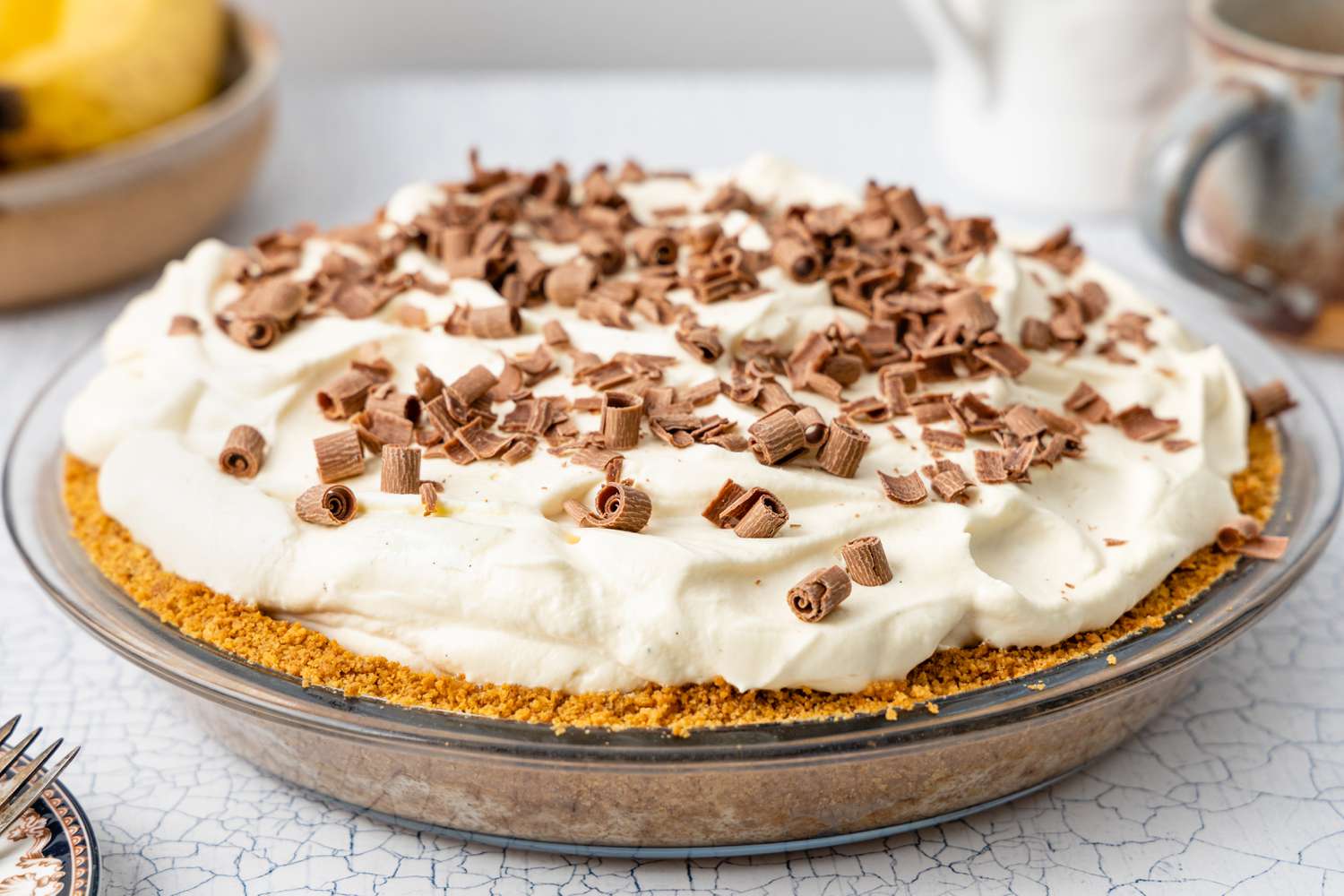 In a Pie Pan, Banoffee Pie Topped with Chocolate Shavings, and in the Surroundings, a Plate with Some Forks, a Bowl of Bananas, and Coffee Mugs