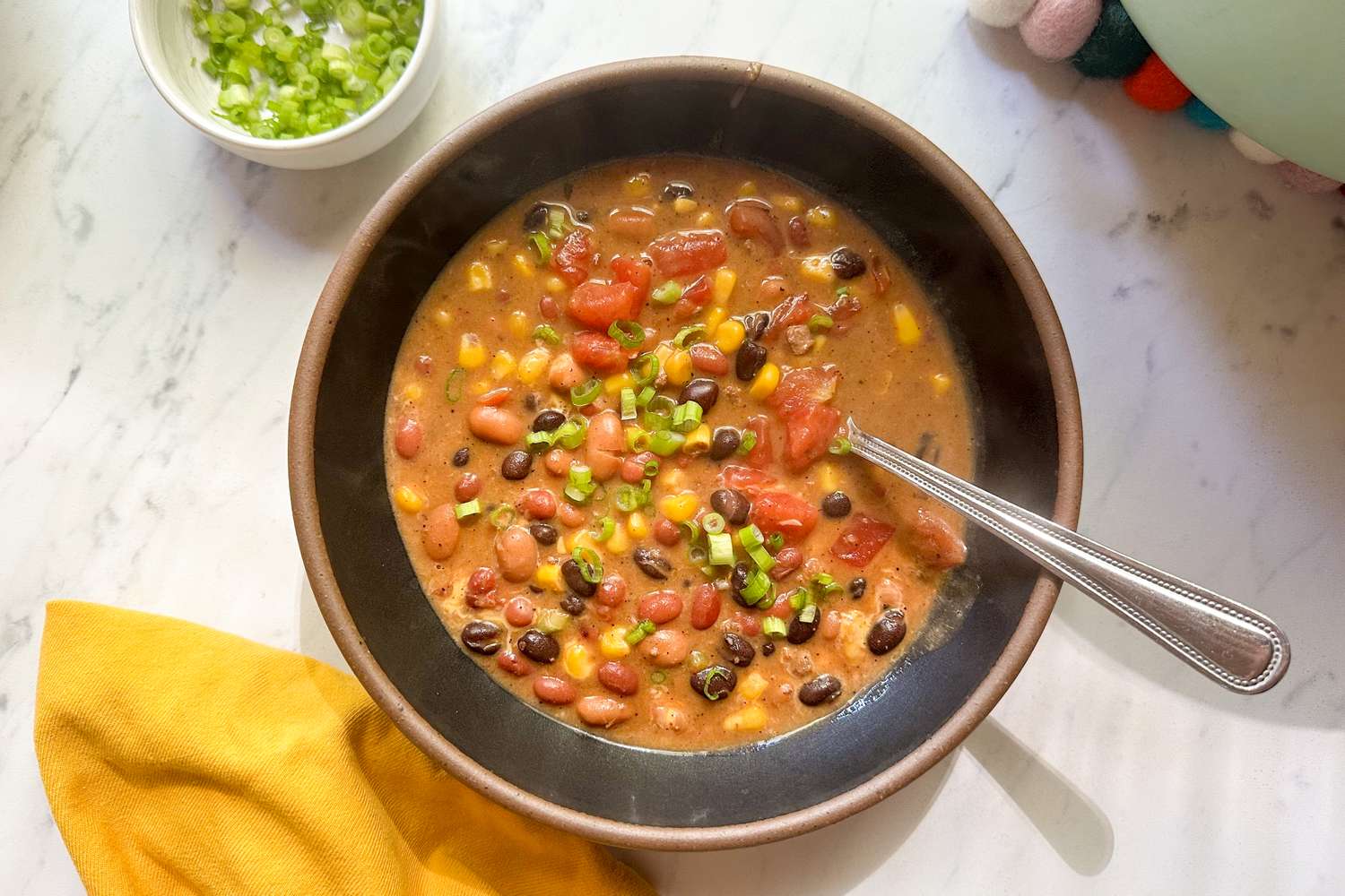A bowl of soup containing beans corn tomatoes and green onions with a spoon and a yellow napkin nearby