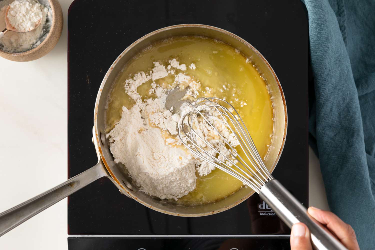 Flour whisked into a pot with melted butter on an portable induction stovetop next a bowl of flour and a blue kitchen towel to a greenish