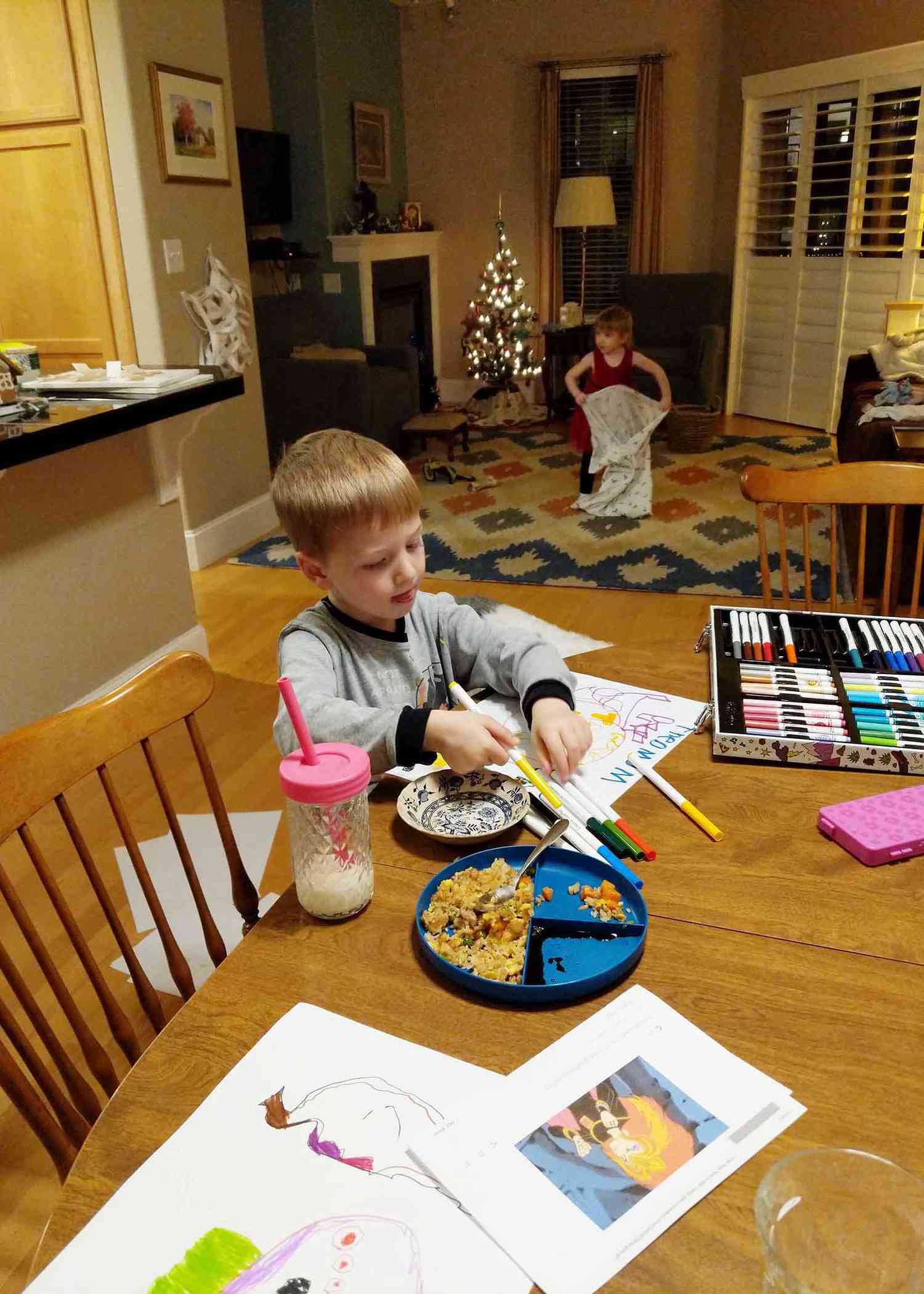 Young boy sitting at a table with pork fried rice with frozen peas and carrots on a plate. Markers and papers are spread on the table while he colors. Little girl is playing in the background with a blanket.