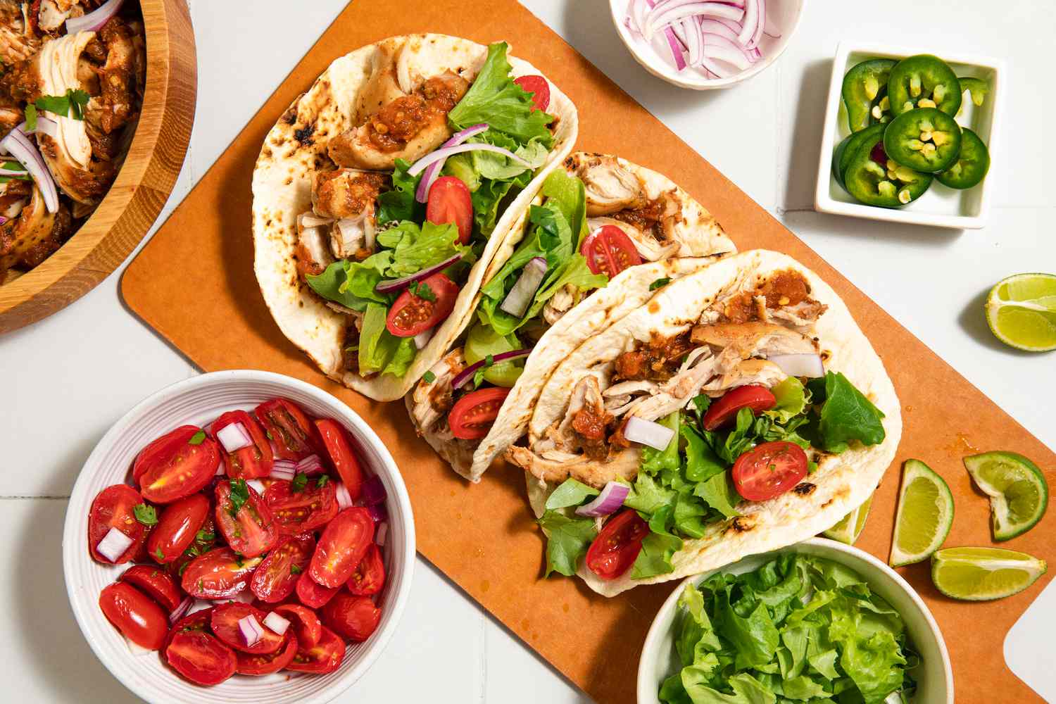 Three chicken tacos with lettuce and tomatoes on a wooden surface next to bowls of toppings and lime wedges