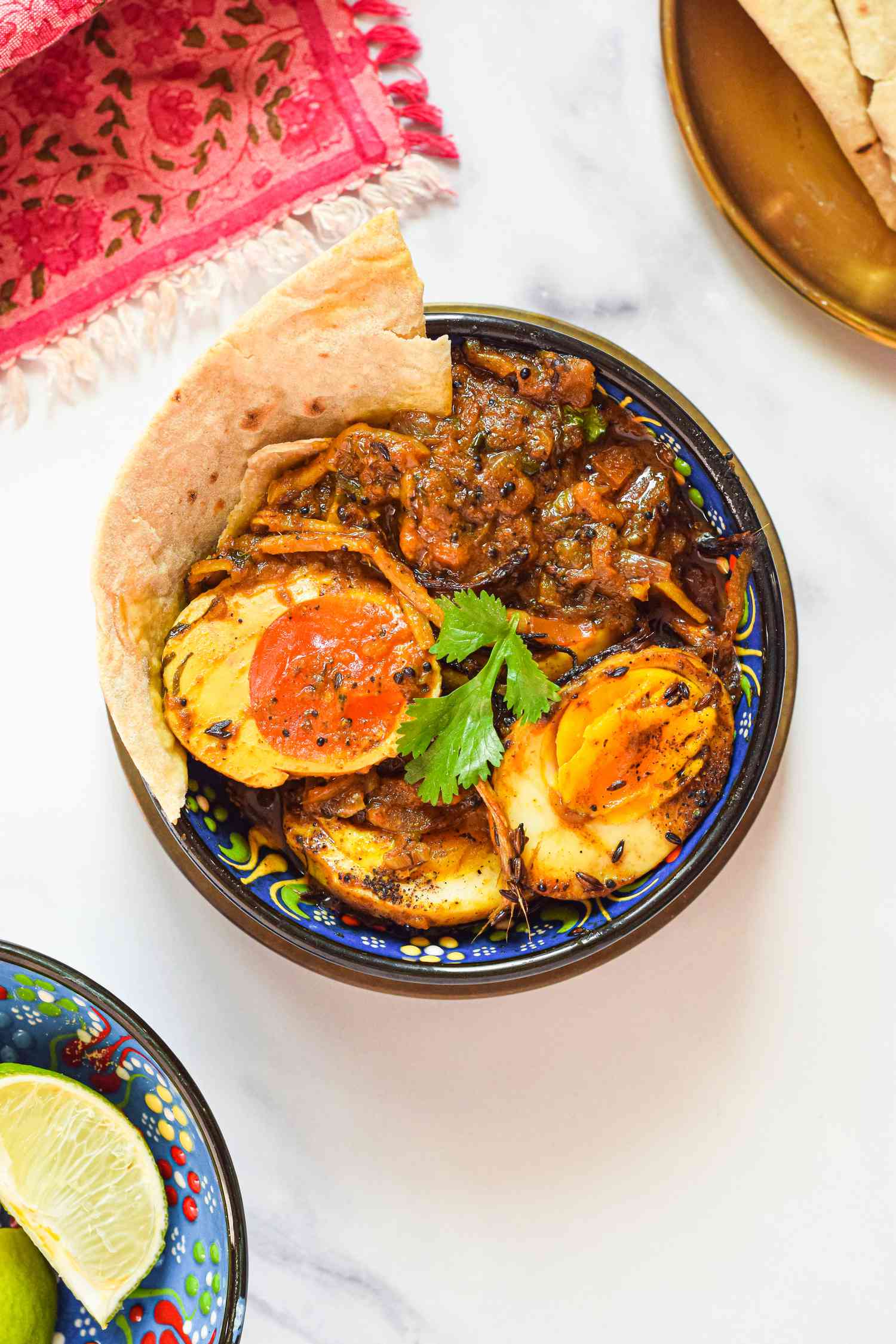 Serving of Egg Masala Curry with Roti in a Bowl Next to a Plate with More Roti and a Bowl with Lime Wedges