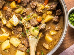 A pot of beef stew containing meat potatoes and carrots with a wooden spoon scooping it up