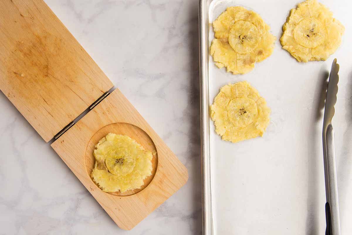 Smashing plantains in a tostonera and setting them on a parchment lined baking sheet.