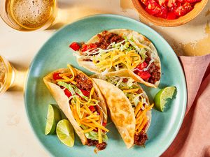 Crispy ground beef tacos on a plate at a table setting with glasses of beer, bowl of diced tomatoes, and a table napkin