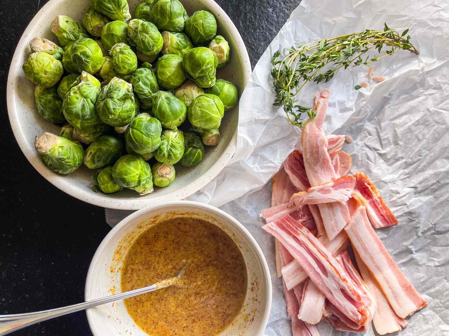 Ingredients for baconwrapped Brussels sprouts including raw Brussels sprouts strips of bacon mustard sauce and fresh thyme