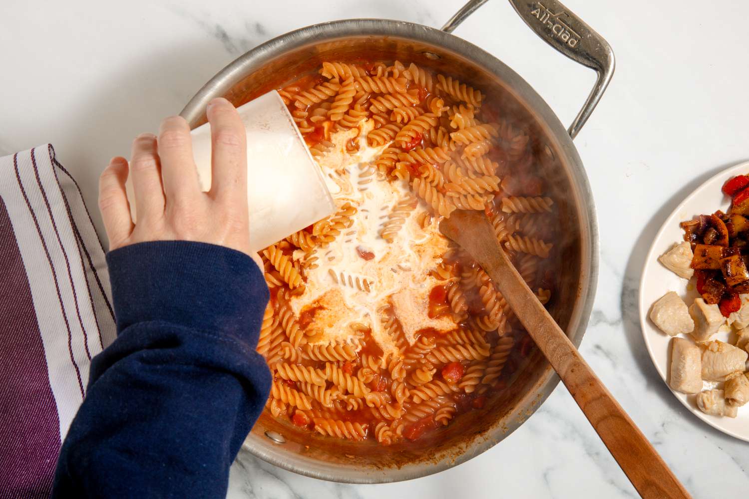 hand pouring heavy cream into pot with tomato and pasta mixture, chicken and veggies on a plate on the side for One-Pan Chicken Fajita Pasta