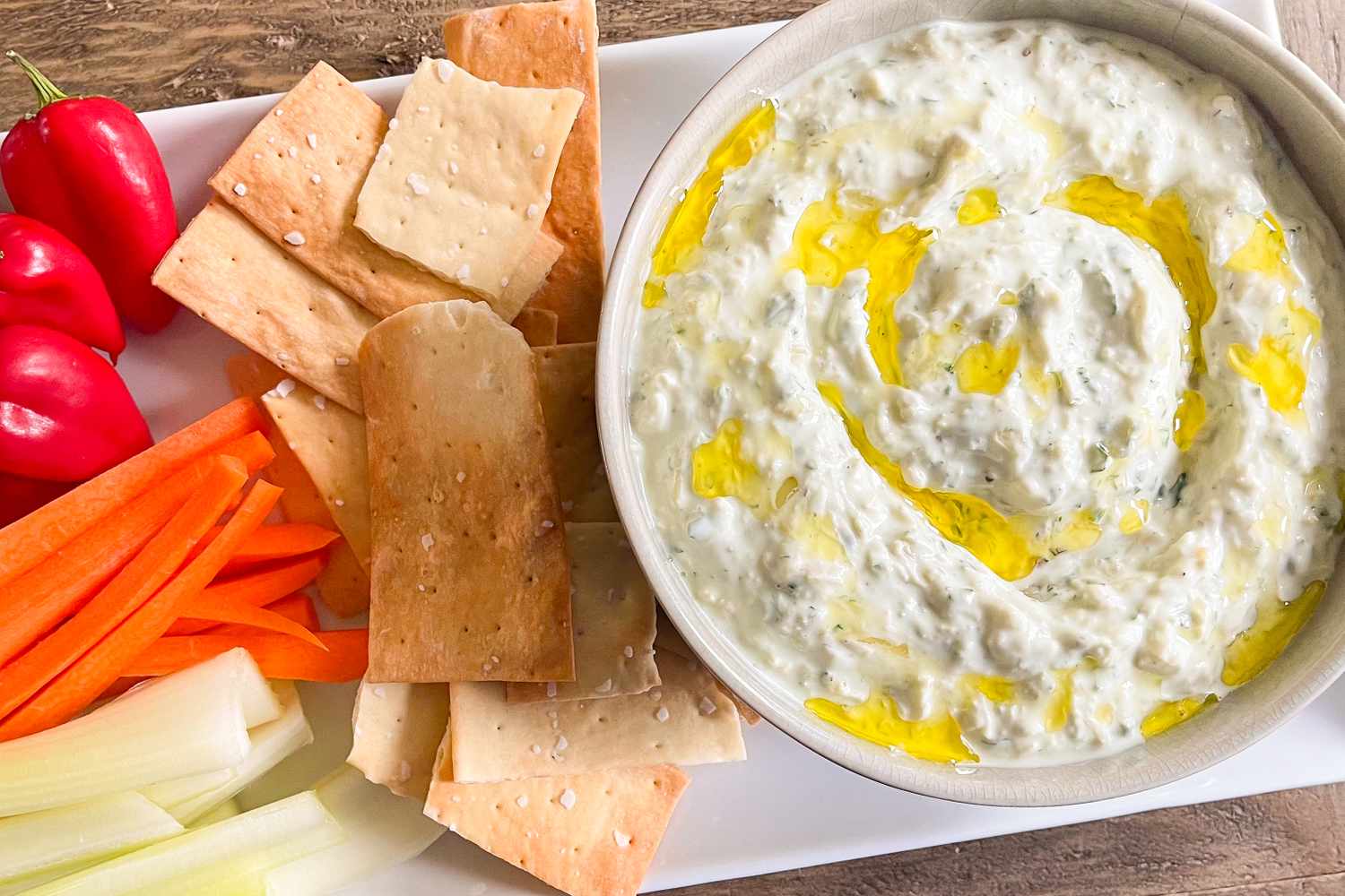 A bowl of dip garnished with oil surrounded by crackers celery carrots and mini peppers on a white platter