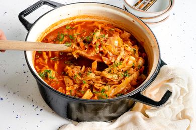 Sweet and Sour Cabbage Soup in a Large Pot with Some Raised Up Using a Wooden Spoon, and Surrounding the Pot, a Kitchen Linen and a Stack of Bowls with Utensils