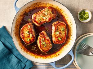 glazed pork chops in a wide skillet at a table setting with a stack of bowls, a table napkin, and a small bowl of sliced scallions