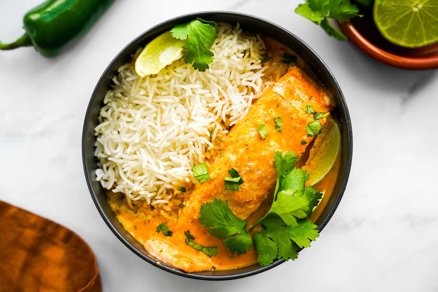 Bowl of Microwave Red Curry Poached Salmon with Rice and Garnished with Cilantro and Lime Wedge, and in the Surroundings, a Kitchen Towel, a Bowl with More Curry, and a Bowl with a Halved Lime and Cilantro