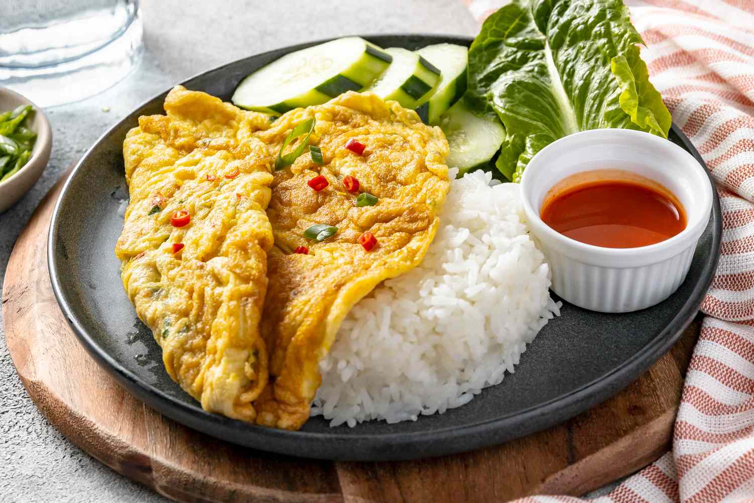 Plate of Kai Jeow (Thai Omelet) with Cucumber Slices, Romaine Leaves, Rice, and a Small Bowl of Sriracha on a Wooden Circular Serving Tray and Next to a Kitchen Towel, a Glass, and a Small Bowl with Toppings