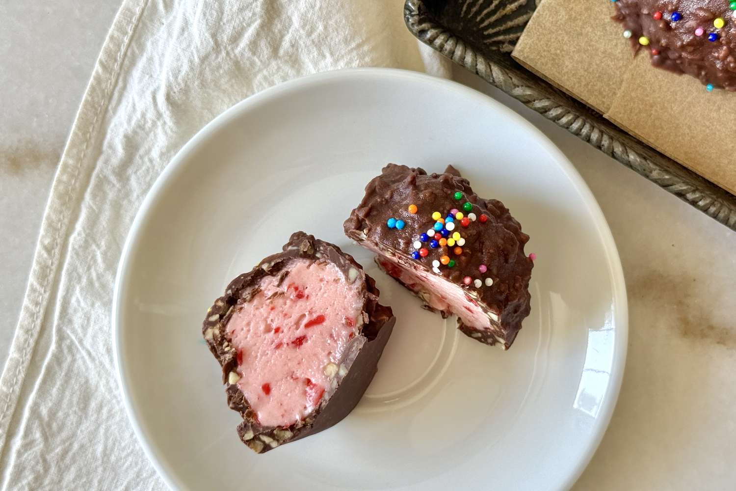 A dessert item with a chocolate coating filled with pink filling and decorated with colorful sprinkles displayed on a white plate
