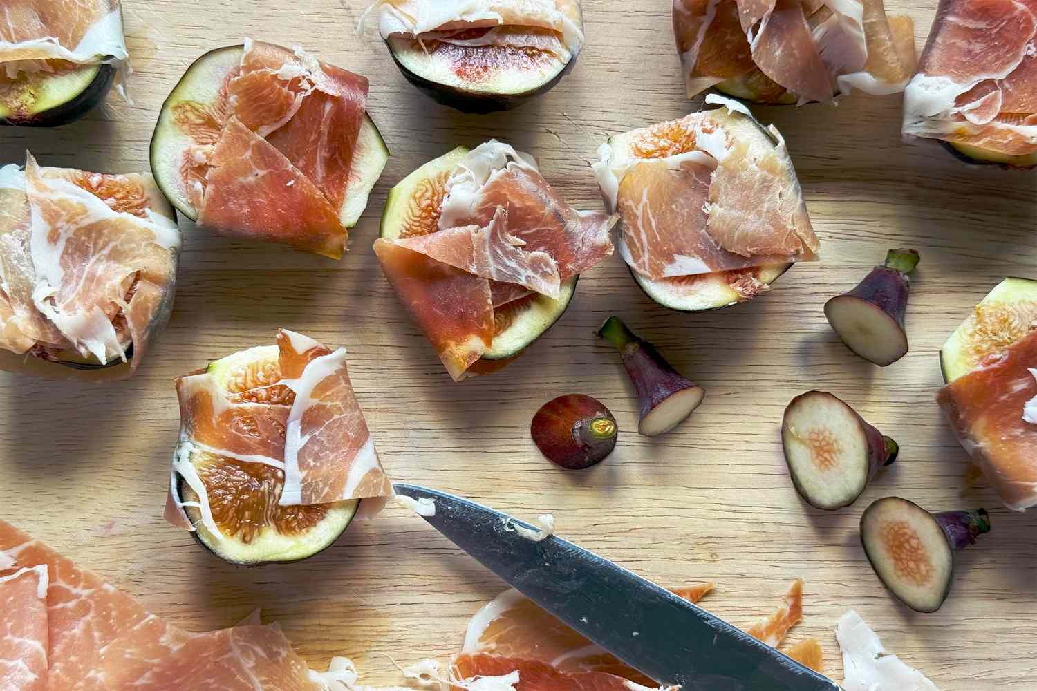 Slices of fig topped with portions of prosciutto on a wooden surface, some whole and cut figs visible