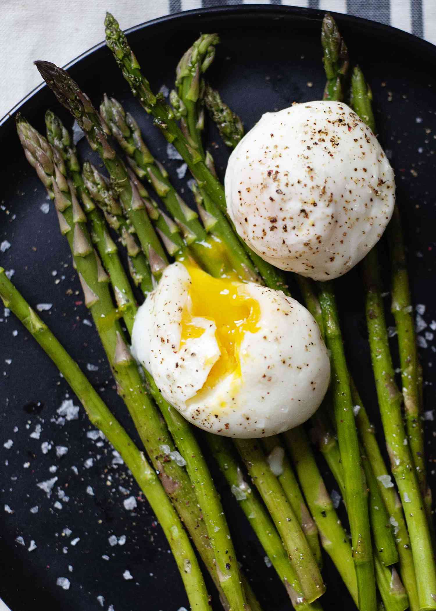 Two simple poached eggs resting on top asparagus. Salt and pepper are sprinkled over both and one egg has a runny yolk visible.