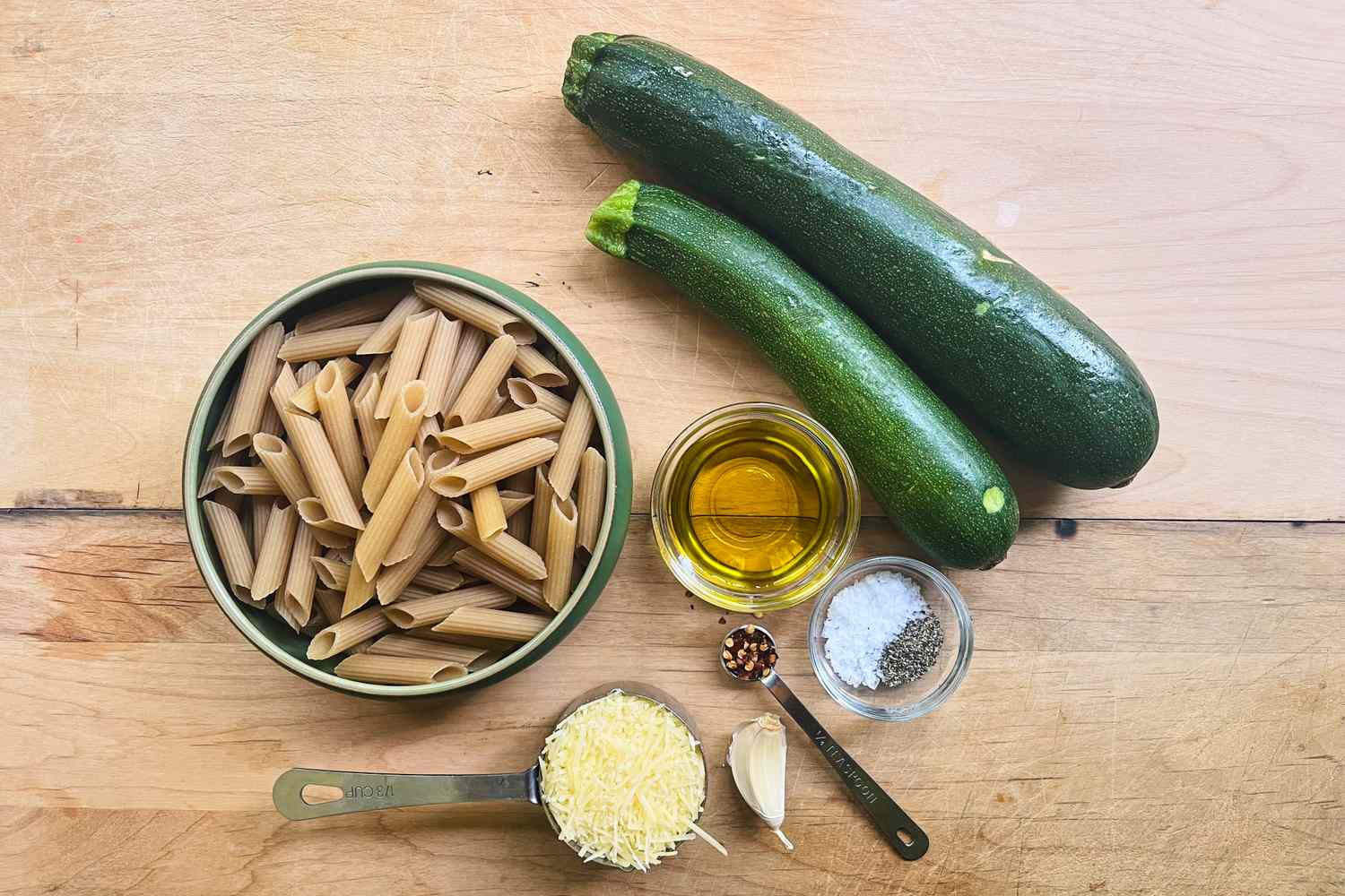 Ingredients on a wooden surface including penne pasta zucchini Parmesan cheese olive oil garlic and spices